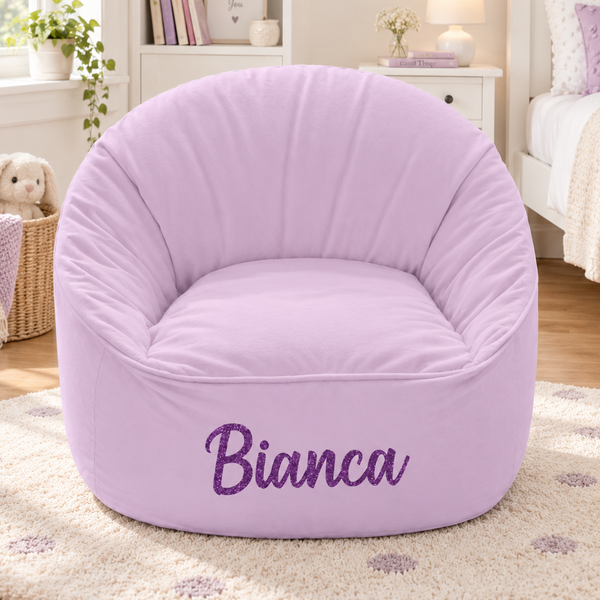 A light purple bean bag chair with the name 'Bianca' embroidered in purple script on the front, set in a cozy, softly lit bedroom.