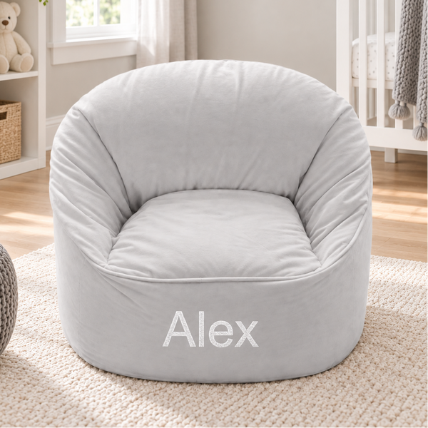 A light gray plush bean bag chair with the name 'Alex' printed in white on the front, set in a styled nursery room next to a crib and window.
