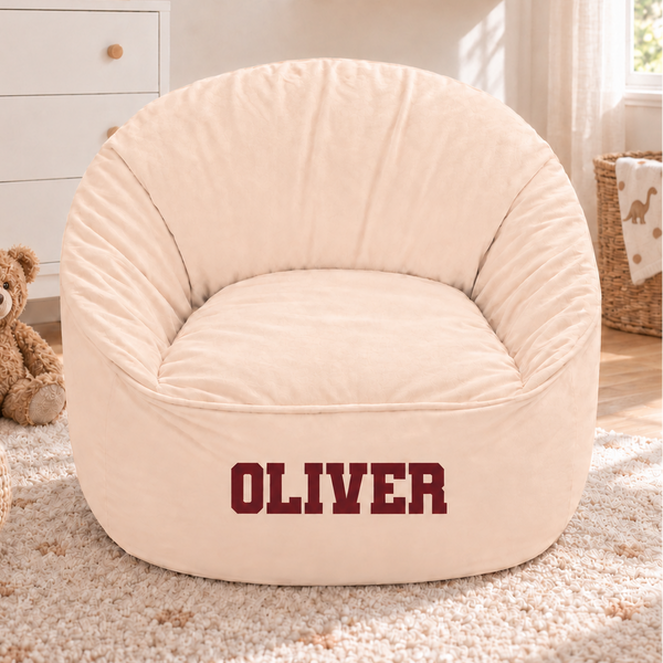 A cream-colored plush bean bag chair with the name 'OLIVER' embroidered in dark red on the front, set in a child's bedroom on a textured rug.