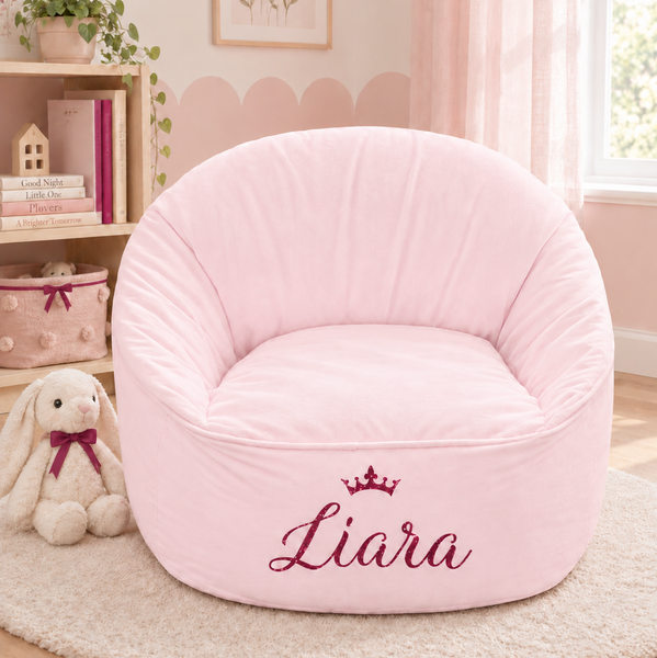 A round, plush pink bean bag chair with personalised name on the front, styled in a child's bedroom setting on a beige rug.