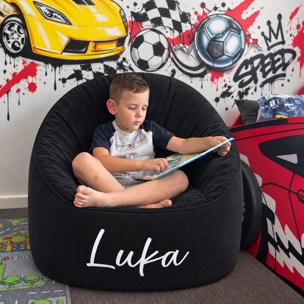 A young boy sits cross-legged in a large black bean bag chair with the name 'Luka' printed in white script on the front. The room has a sports-themed wall mural with a yellow race car, soccer balls, and graffiti-style 'SPEED' text.