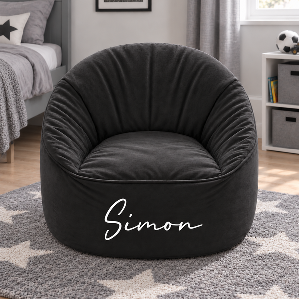 A dark gray plush bean bag armchair with the name 'Simon' printed in white script on the front, set in a modern bedroom on a gray star-patterned rug.